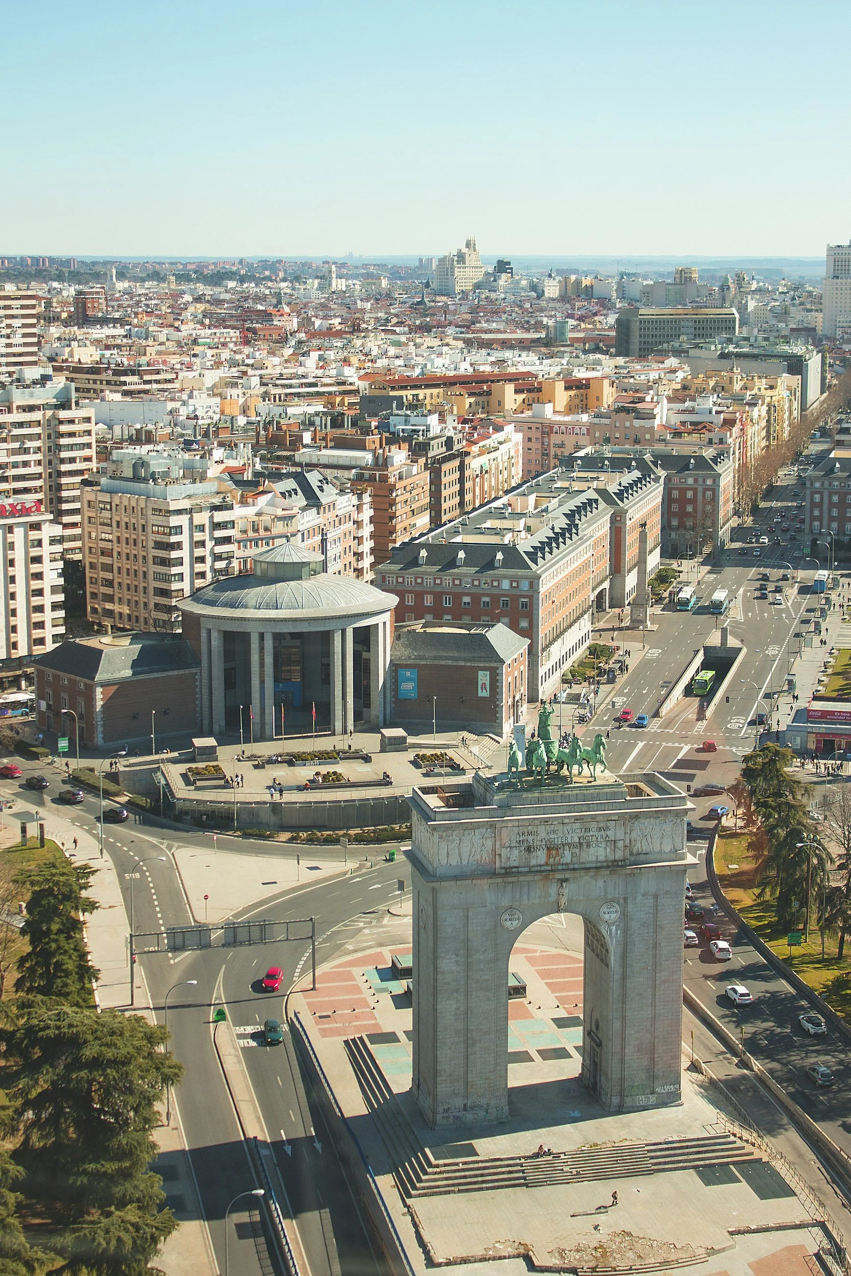 An aerial view of a cityscape featuring a historic arch monument with equestrian statues on top, surrounded by roads, cars, and modern buildings in the background under a clear blue sky.