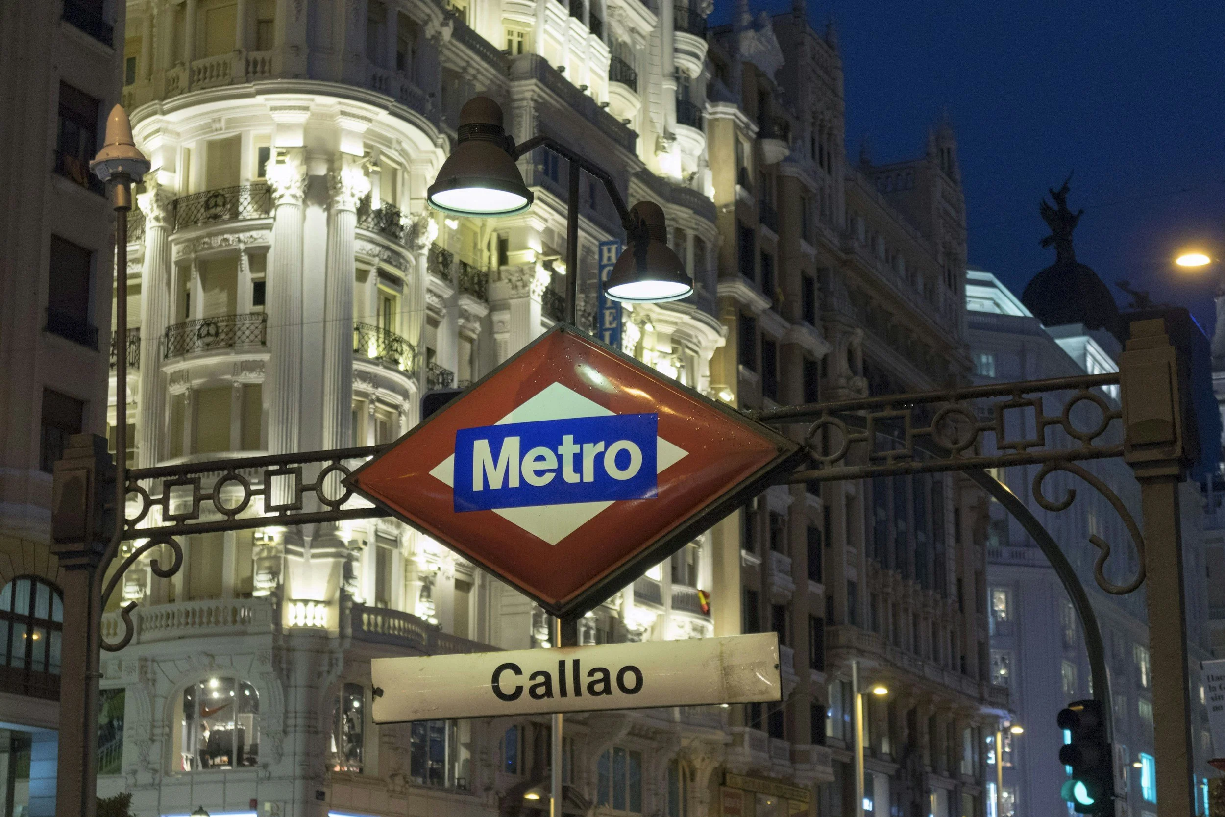Night view of Madrid metro sign at Calle Callao with illuminated historic buildings and street lamps in the background.