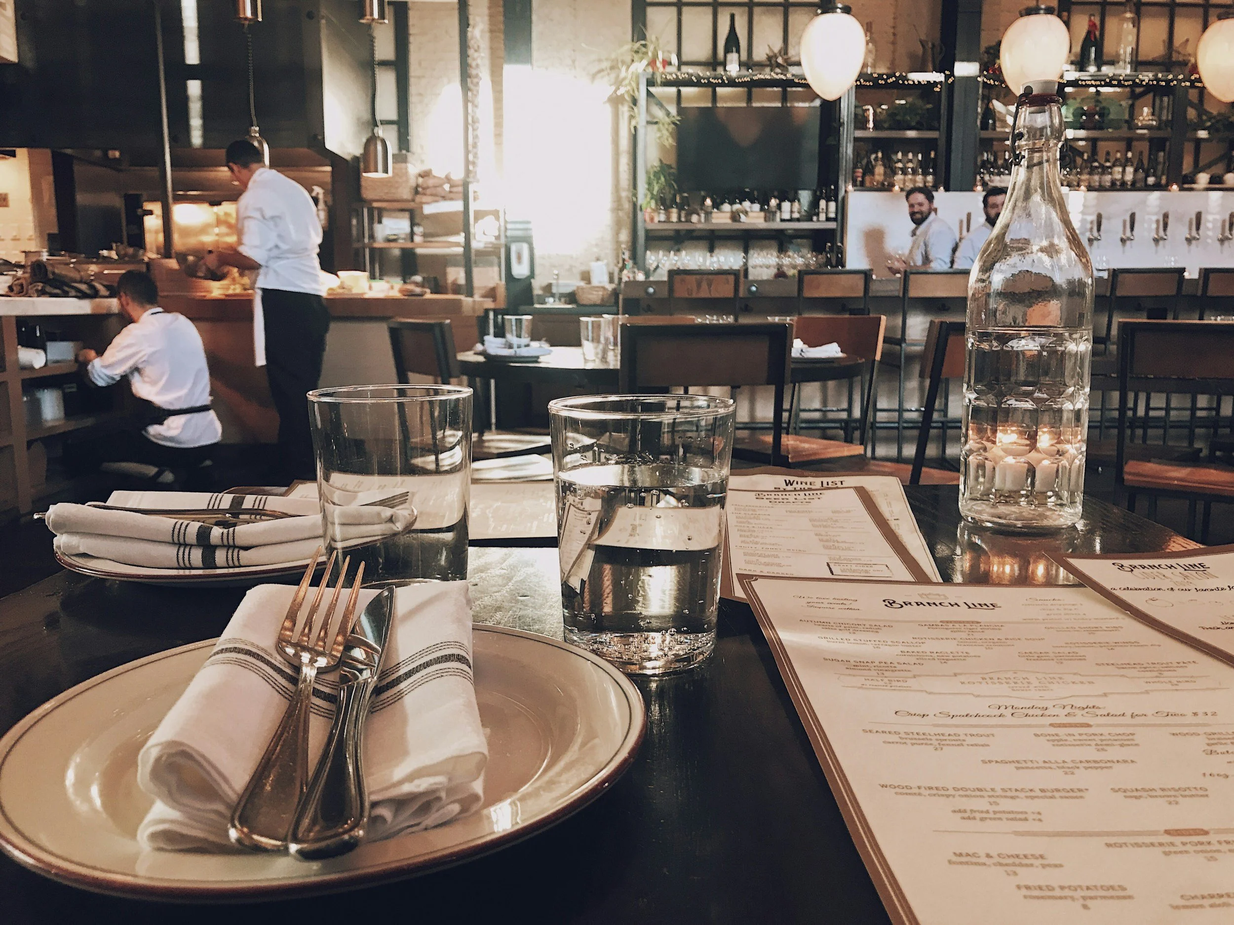 A restaurant table set with a plate, fork, and knife on a napkin, a glass of water, and a menu. There are two glasses of water and a bottle of water on the table. The background shows a restaurant interior with servers working and dining tables, warm lighting, and a bar area.