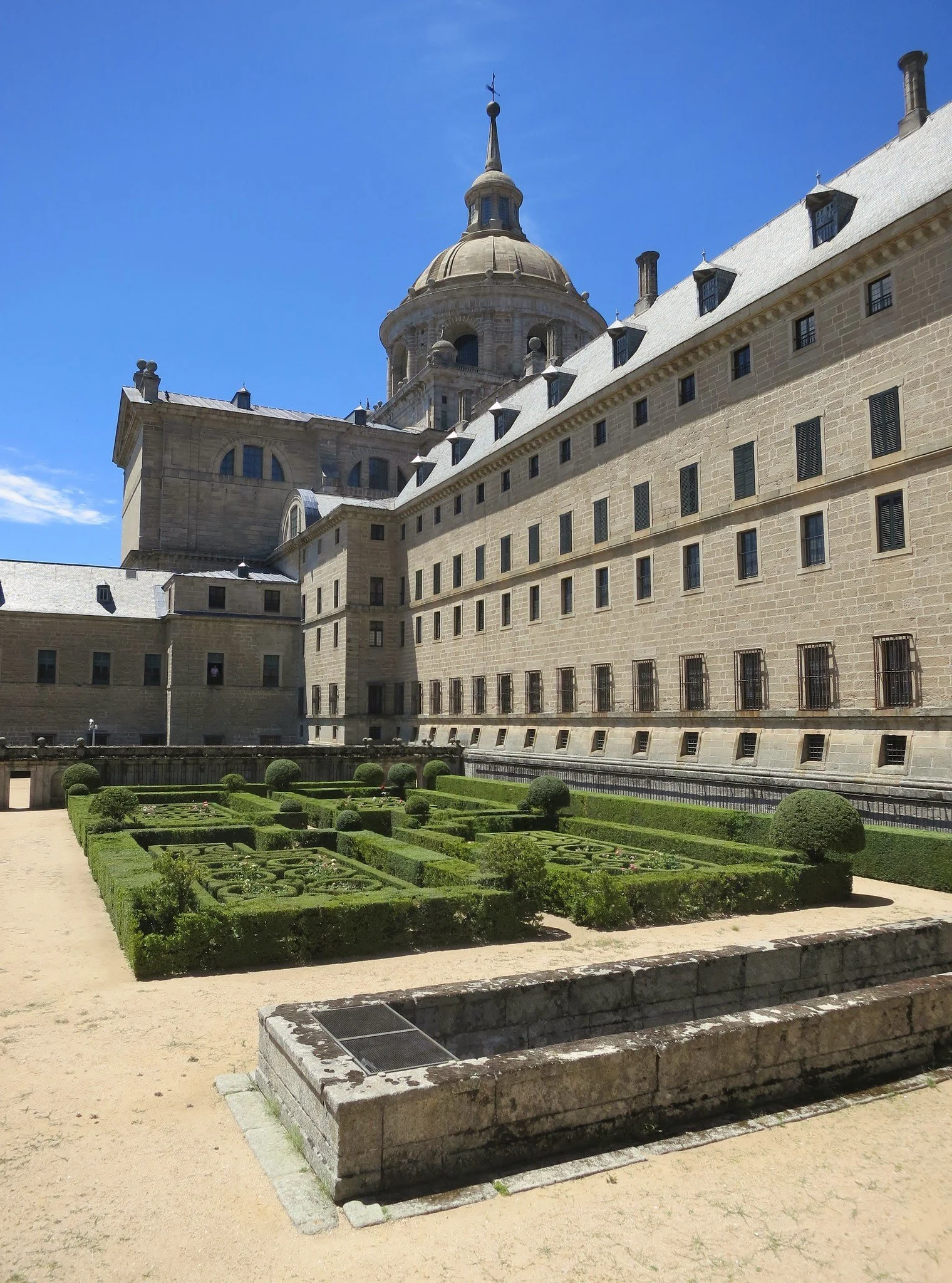 View of a historic European building with a large dome, surrounded by a manicured garden with geometric patterns, bushes, and gravel paths, under a clear blue sky.