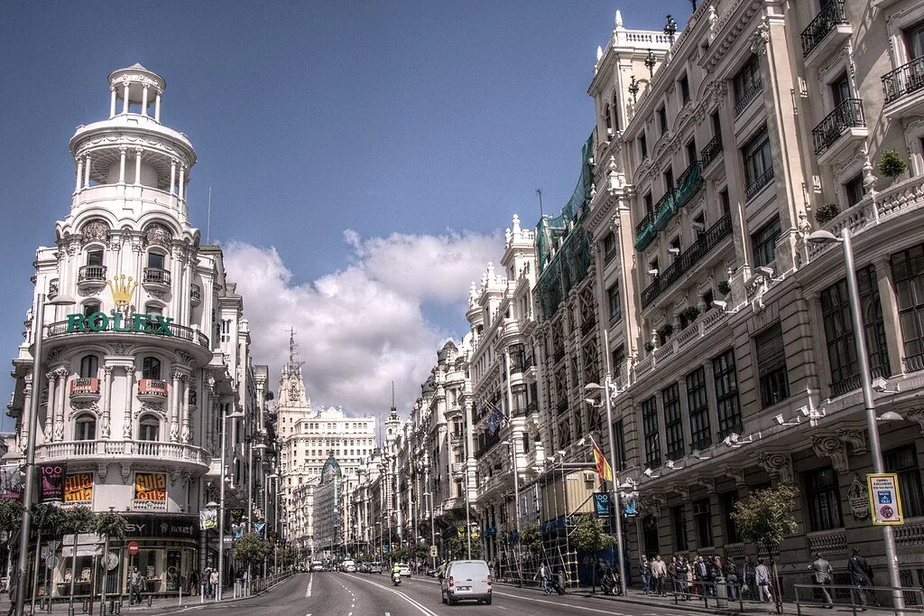 City street with ornate white buildings, people walking, and a few cars under a partly cloudy sky.