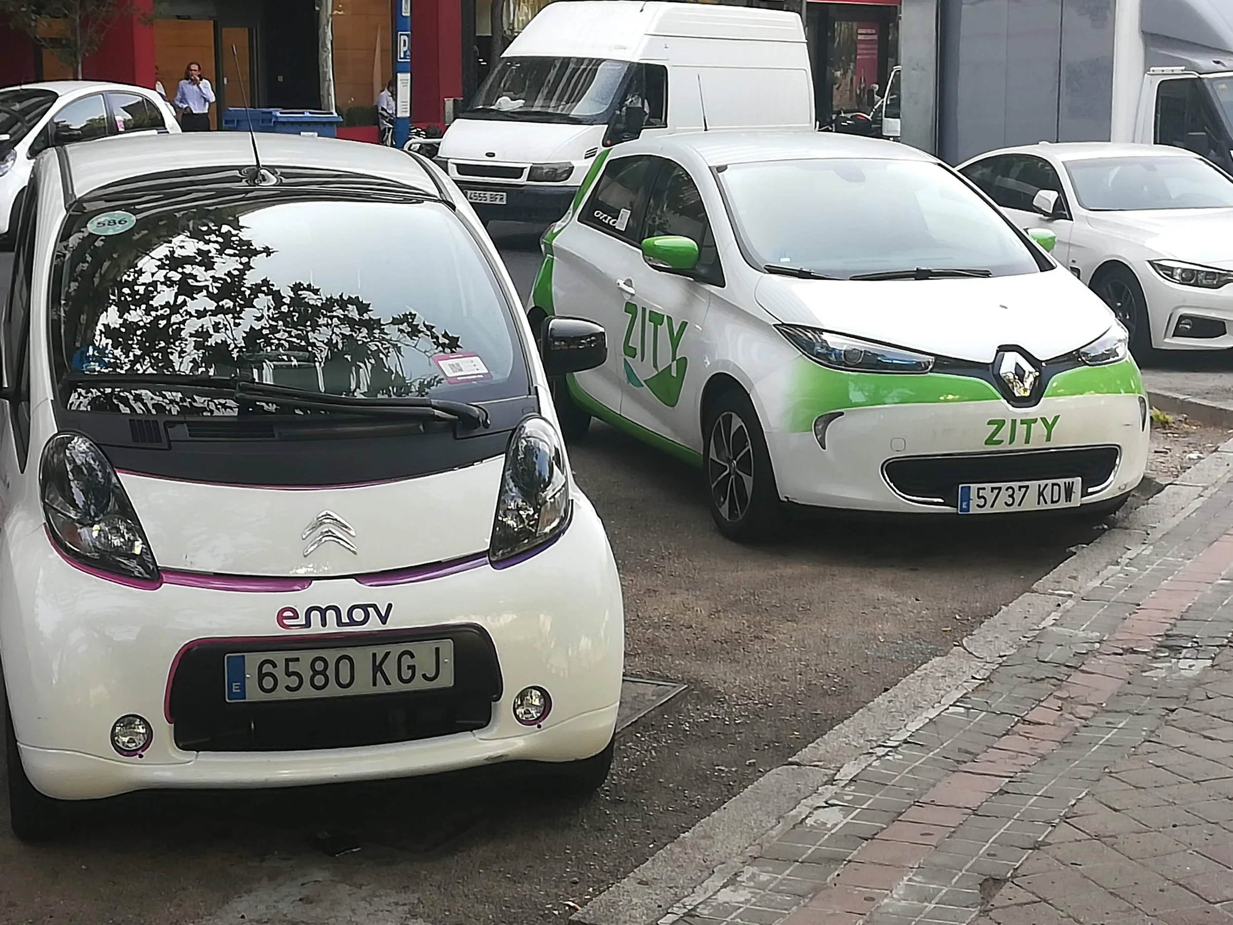 Street parking with several small electric cars, including a white Citroën with 'emov' logo and a white Renault Zity car with green accents, alongside other vehicles and pedestrians in the background.