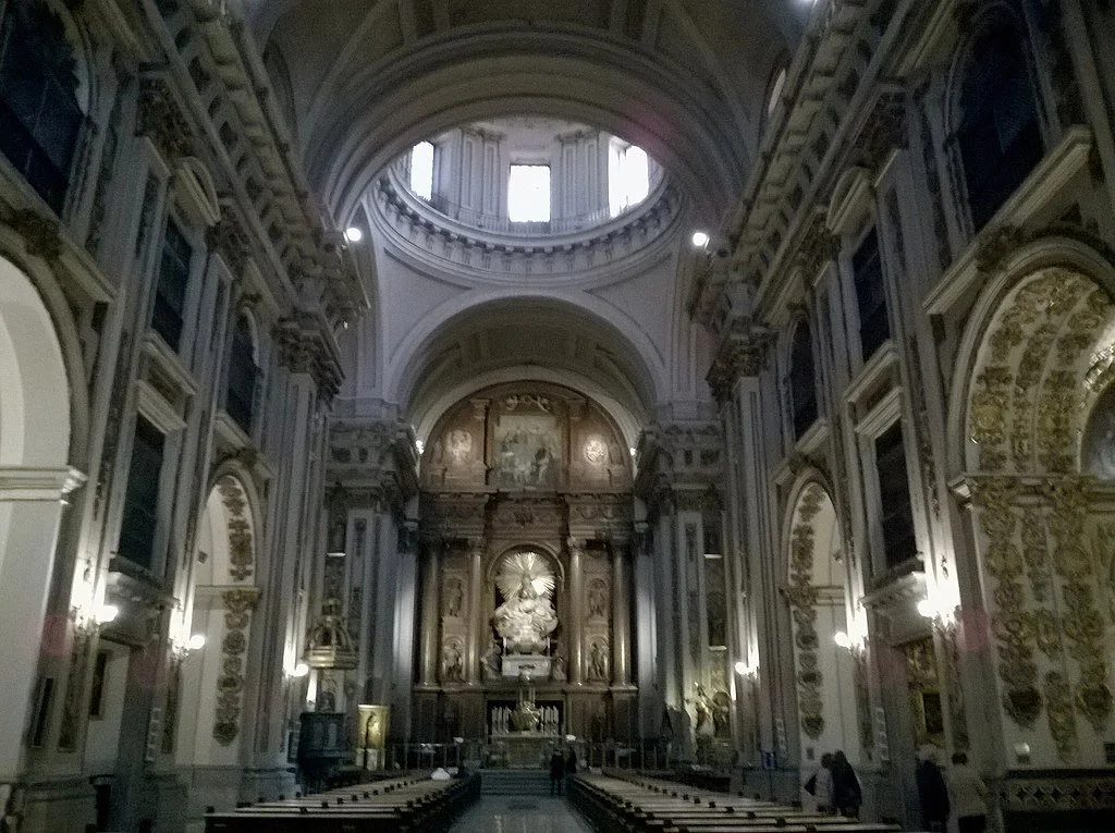 The interior of a grand, historic church with ornate gold and white decorations, high vaulted ceilings, and a large altar at the front.
