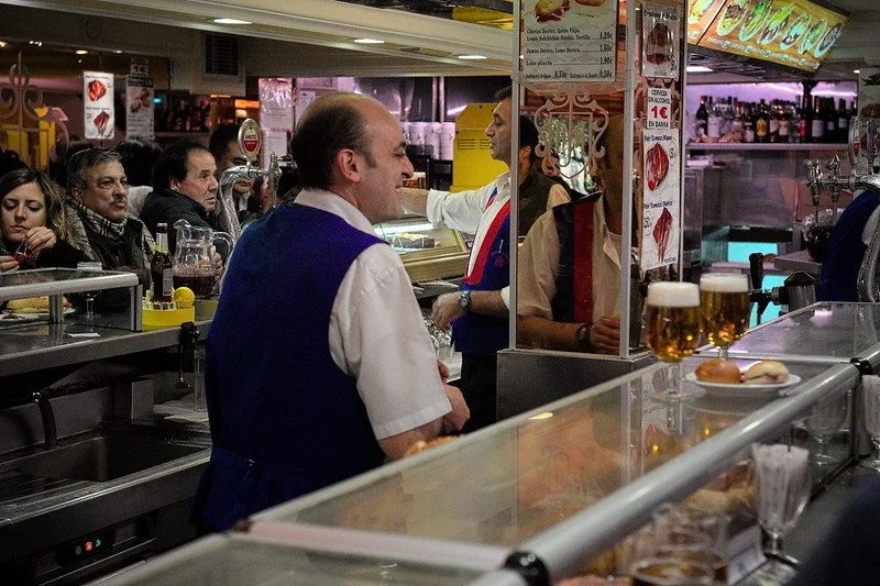 A busy bar or tavern with a crowd of patrons, a bartender, and various drinks on the counter.