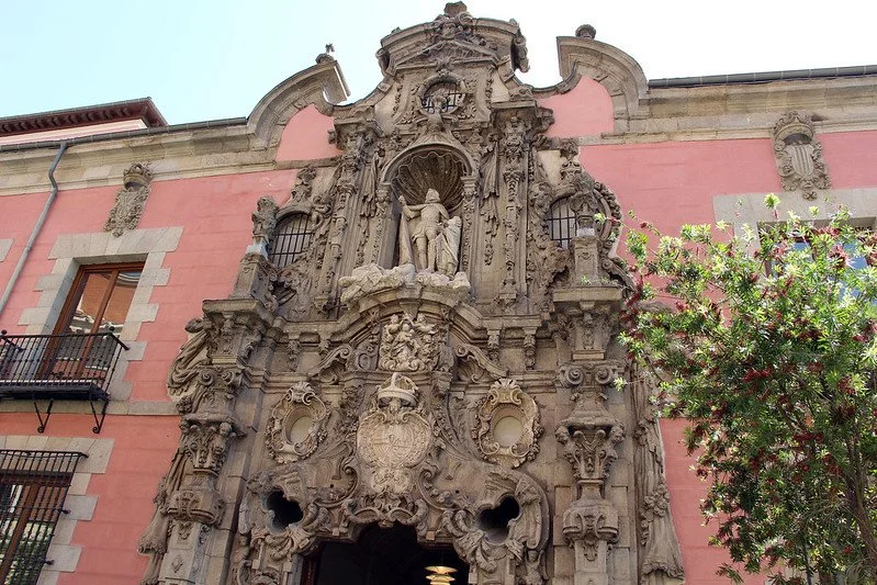 Baroque-style facade of a historic building with intricate stone carvings, statues, and a pink exterior wall, with windows and a small balcony.