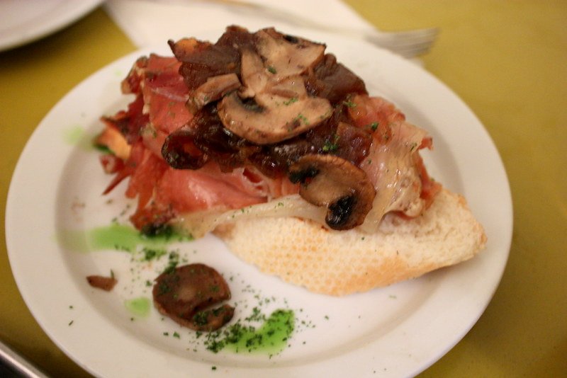 Open-faced sandwich with mushrooms, tomatoes, and ham on a slice of bread, served on a white plate with green herbs and a black olive.