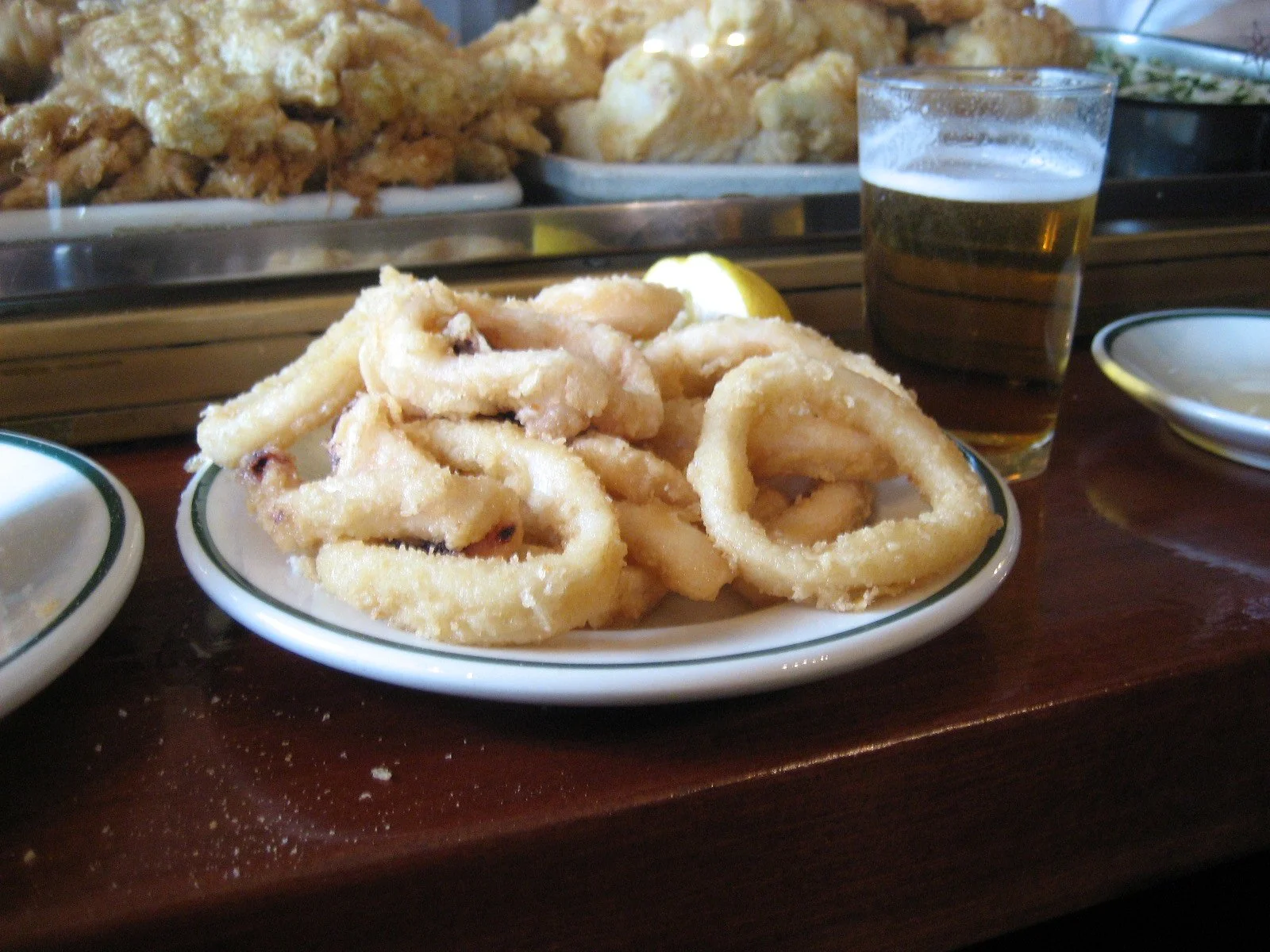 Fried calamari on a white plate with a green rim, served with lemon wedge and a glass of beer on a wooden table in a restaurant.