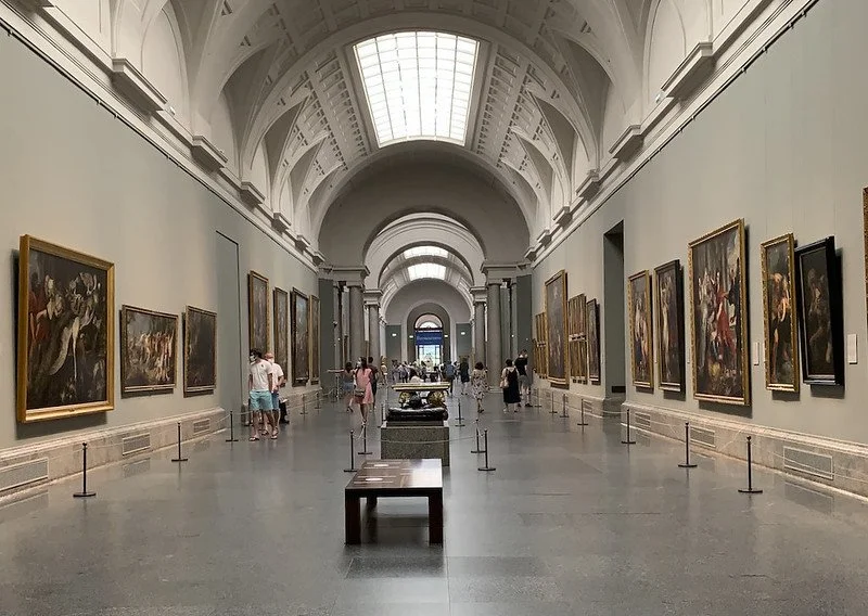 Art gallery with large paintings on the walls, visitors walking and observing the artwork, in a spacious hall with high arched ceilings and skylights.