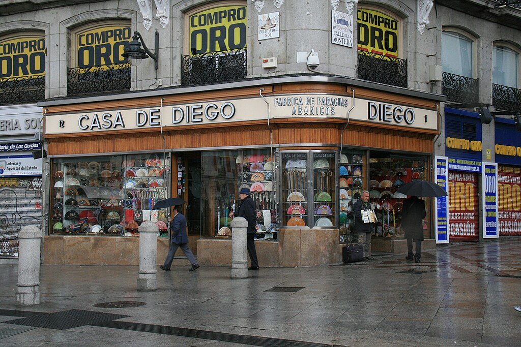 A storefront named Casa de Diego selling umbrellas and hats on a rainy day, with people walking with umbrellas in front.