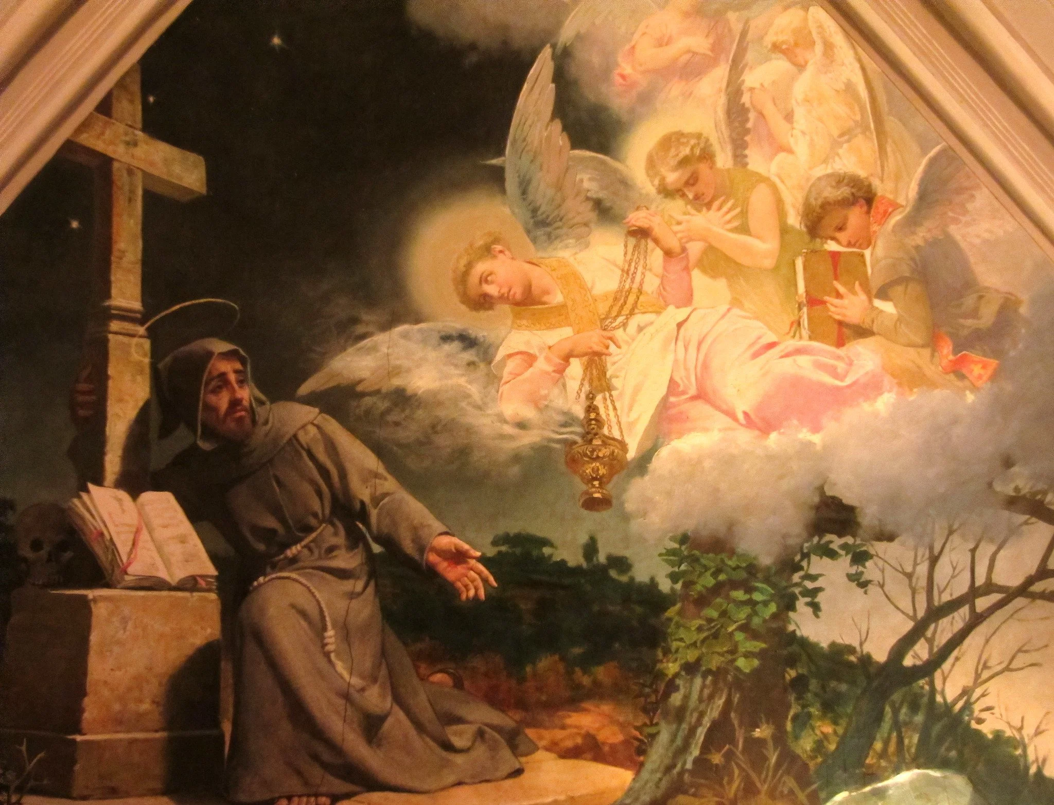 A religious scene with a statue of a man in brown robes praying near a cross, with a book open nearby. Behind him is a painting or mural depicting angels in the sky, one holding a lantern, and others holding a gift, with clouds and a heavenly background.