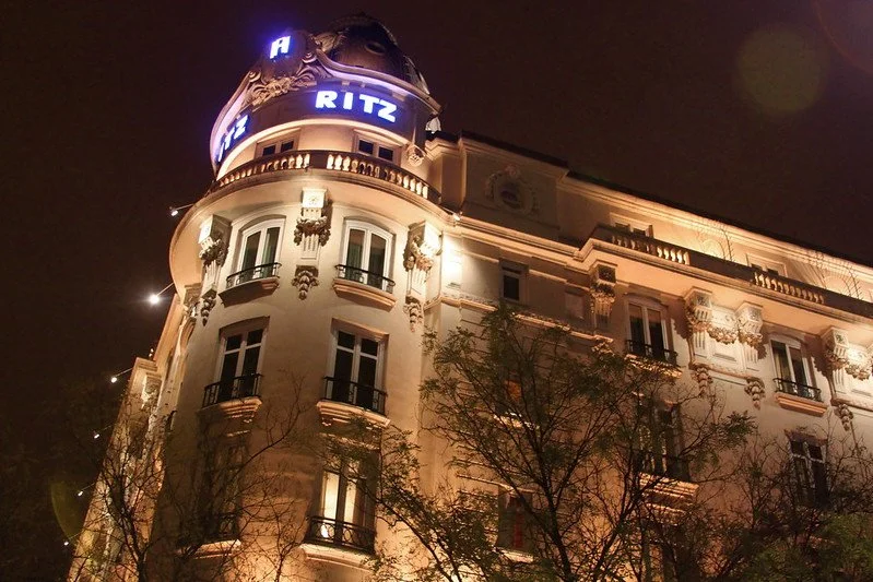 Night view of a historic, elegant hotel building with ornate architectural details, illuminated signage reading "RITZ," and trees in the foreground.