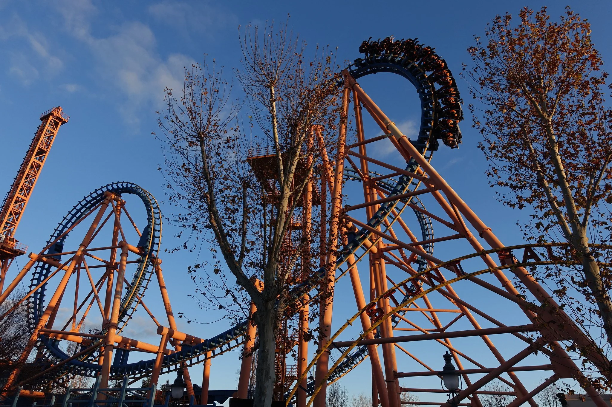 A roller coaster with a blue track and orange supports viewed against a blue sky with some clouds. Some leafless trees are in the foreground.