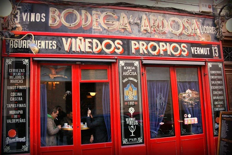 Exterior of a Spanish bodega named 'Bodega Ardosá' with a red door, window panels, and black chalkboard signs listing beverages and food items.