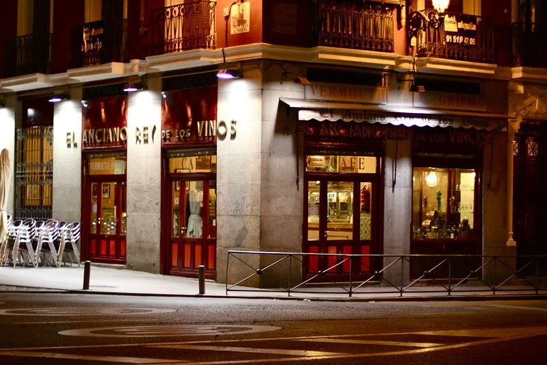 Night view of a wine shop with red doors and illuminated signs, some chairs stacked outside, on a city street.