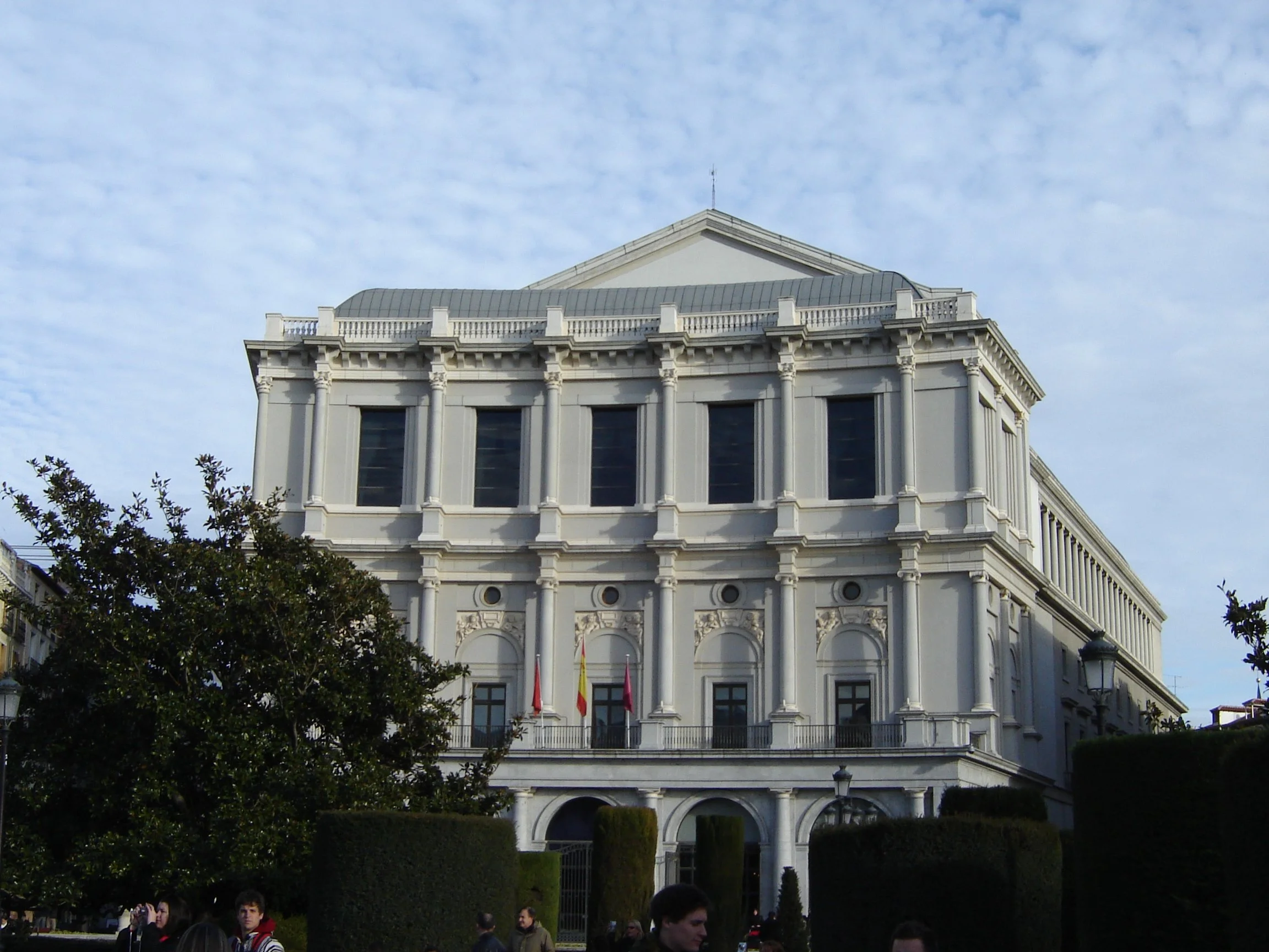 A large, white, neoclassical building with columns, arched windows, and a balcony with flags, under a partly cloudy sky.