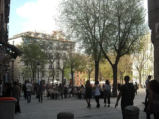 Crowded urban outdoor scene with people walking and sitting at tables, tall trees, and buildings in the background during daytime.