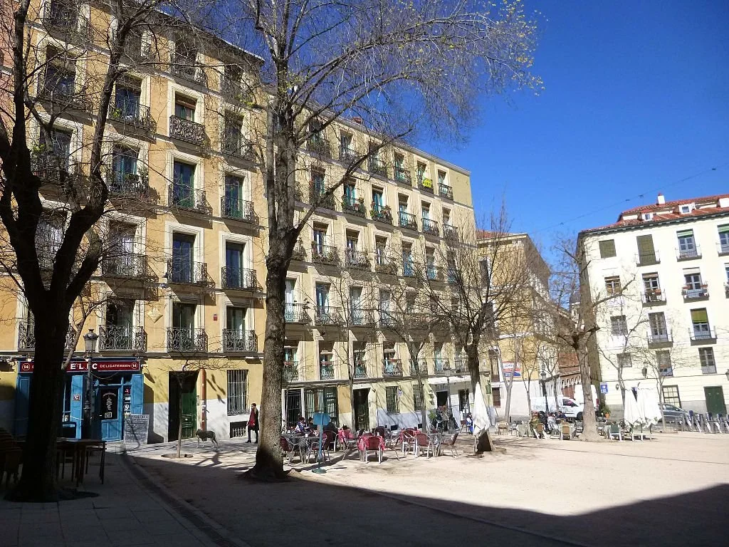 A street scene with apartment buildings, leafless trees, outdoor seating, and a clear blue sky.