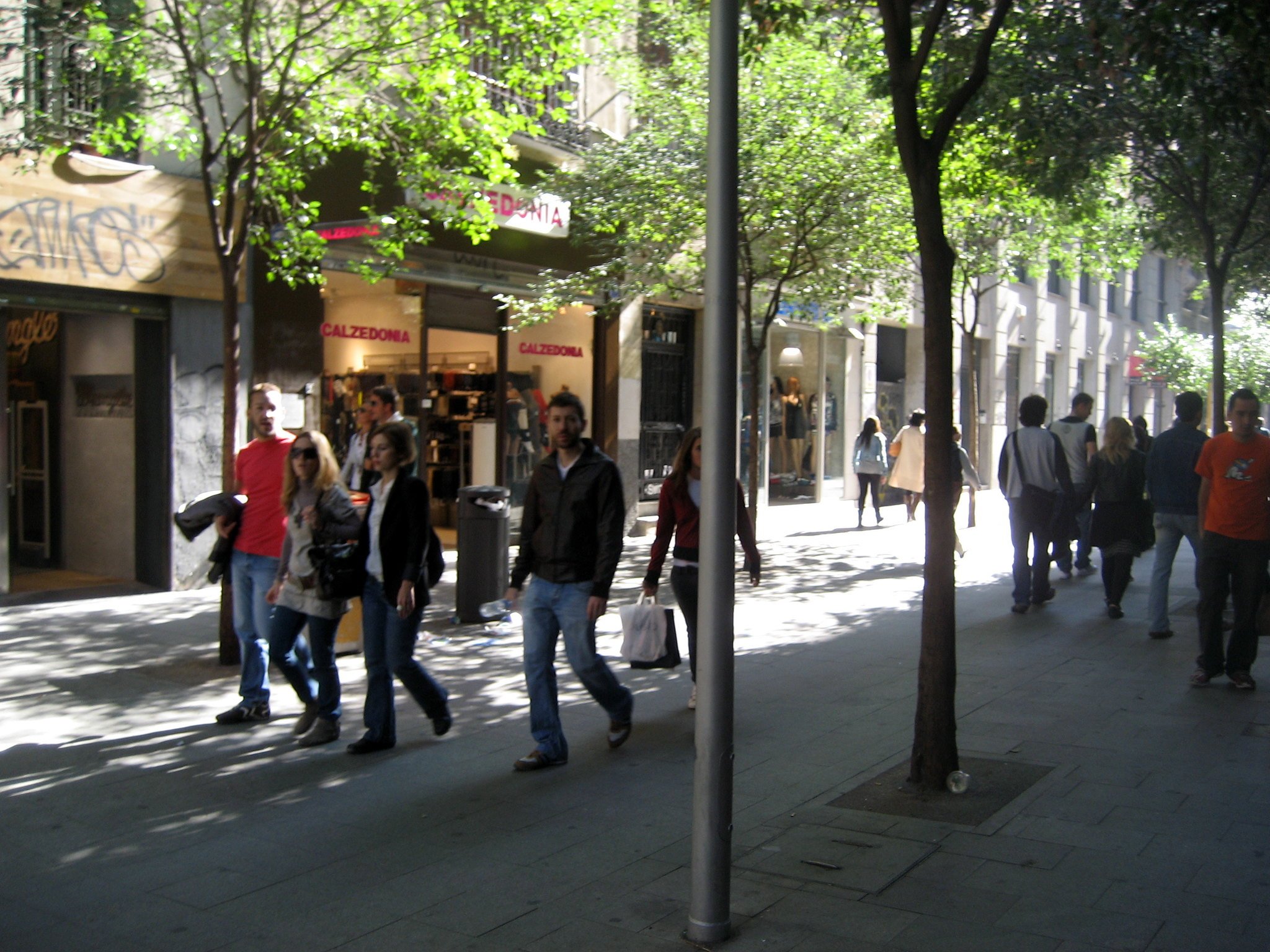 People walking on a city sidewalk lined with trees and storefronts, including a Calzedonia store.