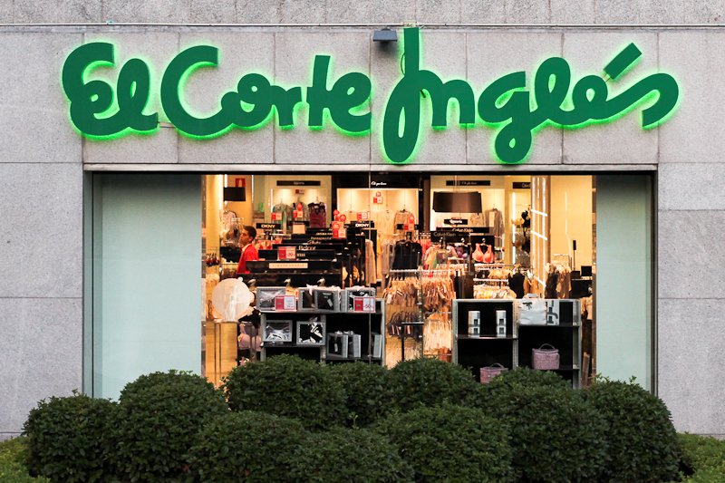 Storefront of The Children's Place retail store with green illuminated sign and items displayed inside, seen through large glass window, with neatly trimmed bushes in front.