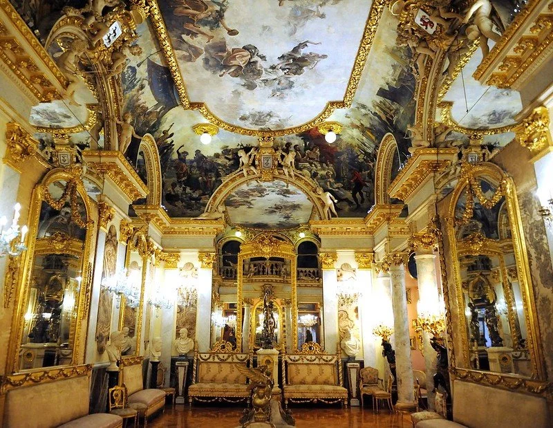 A luxurious, ornate room with gold accents, classical sculptures, and an elaborate painted ceiling depicting a mythological scene.