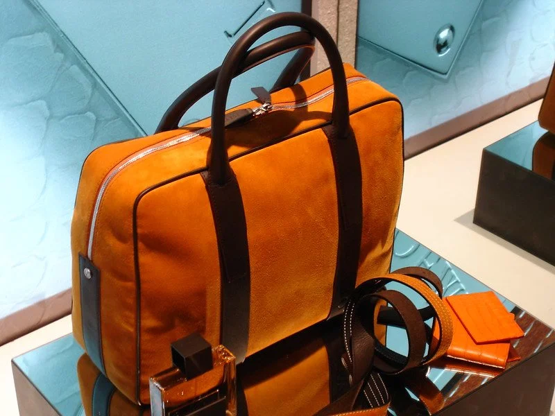 A brown and orange leather briefcase with black handles, accompanied by a matching orange wallet, a brown belt, and a small bottle of cologne, placed on a reflective surface.