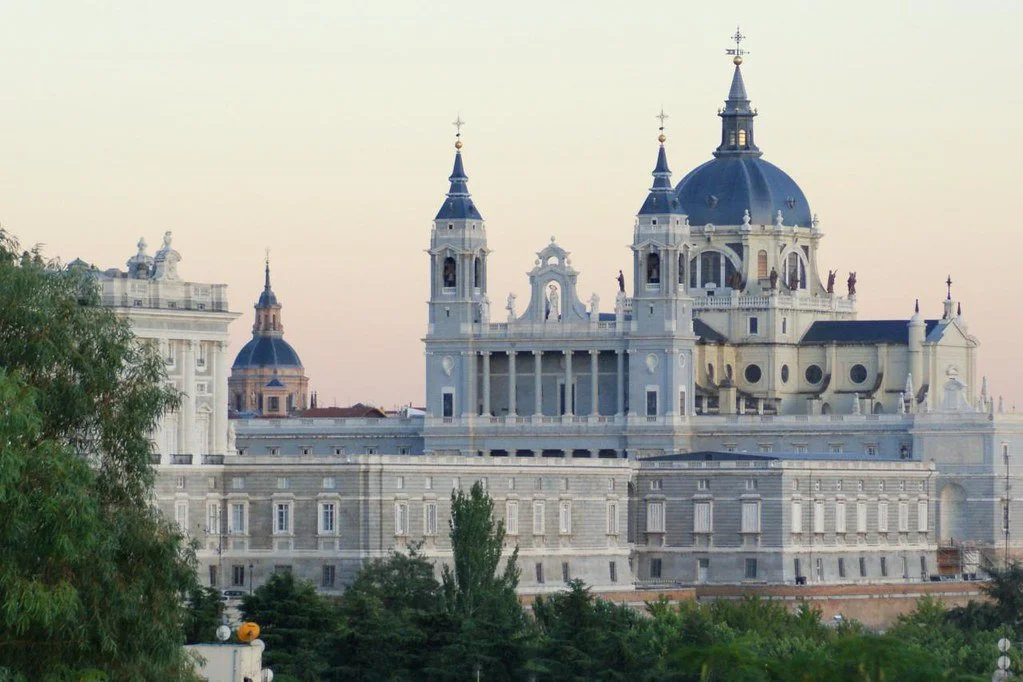 A large, historic cathedral with domes and spires, surrounded by trees, and set against a pale sky.