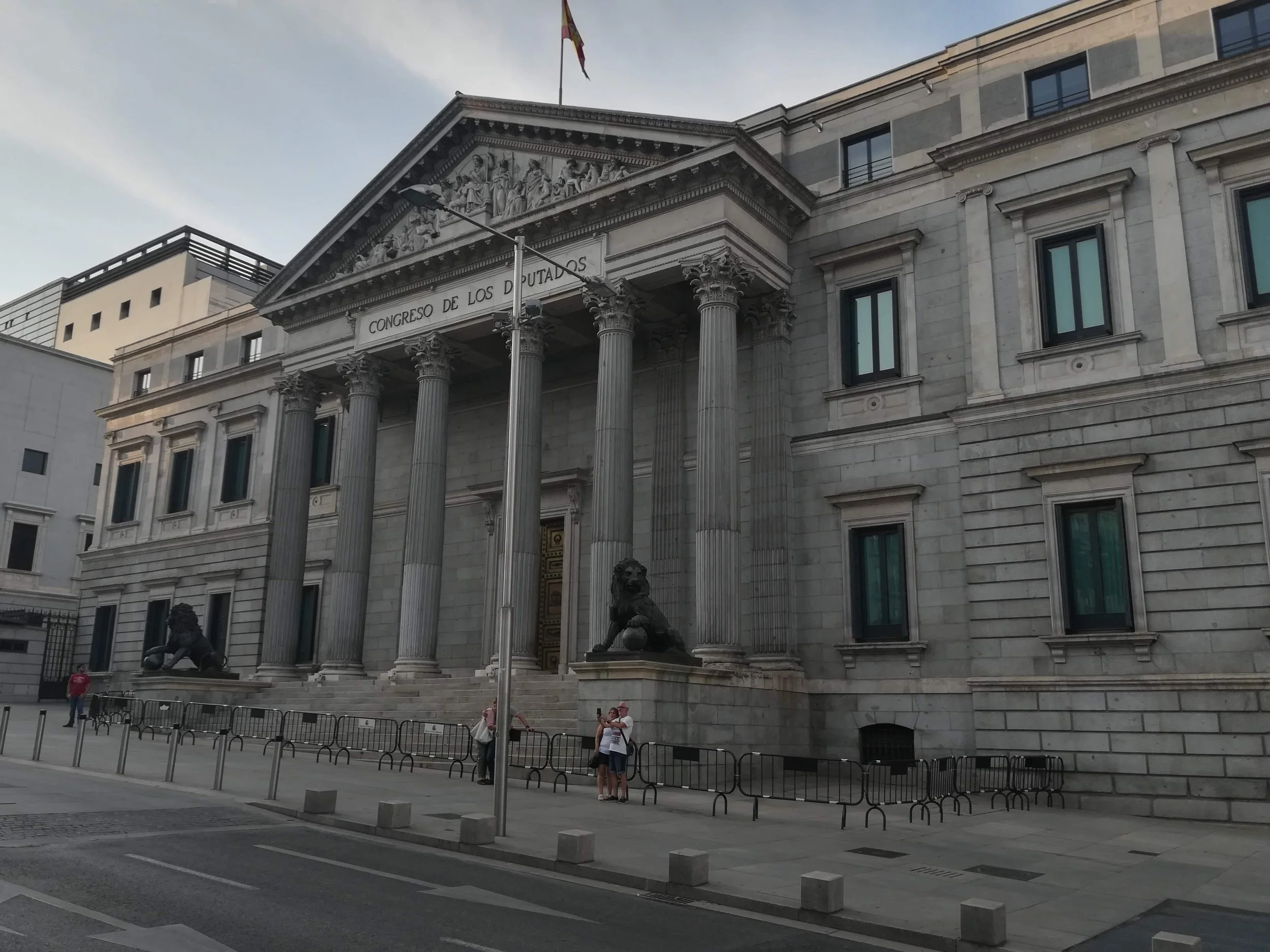 The building is a grand government structure with a classical design, featuring tall columns, statues of lions at the entrance, and a Latin inscription that reads 'Congreso de los Diputados'. There is a flag flying above the building, and a few people are standing near the entrance.