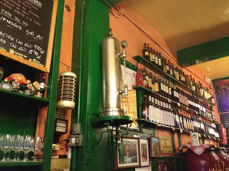 Interior of a restaurant with a large stainless steel distillation bulb, shelves filled with wine bottles, framed pictures, and a chalkboard menu.