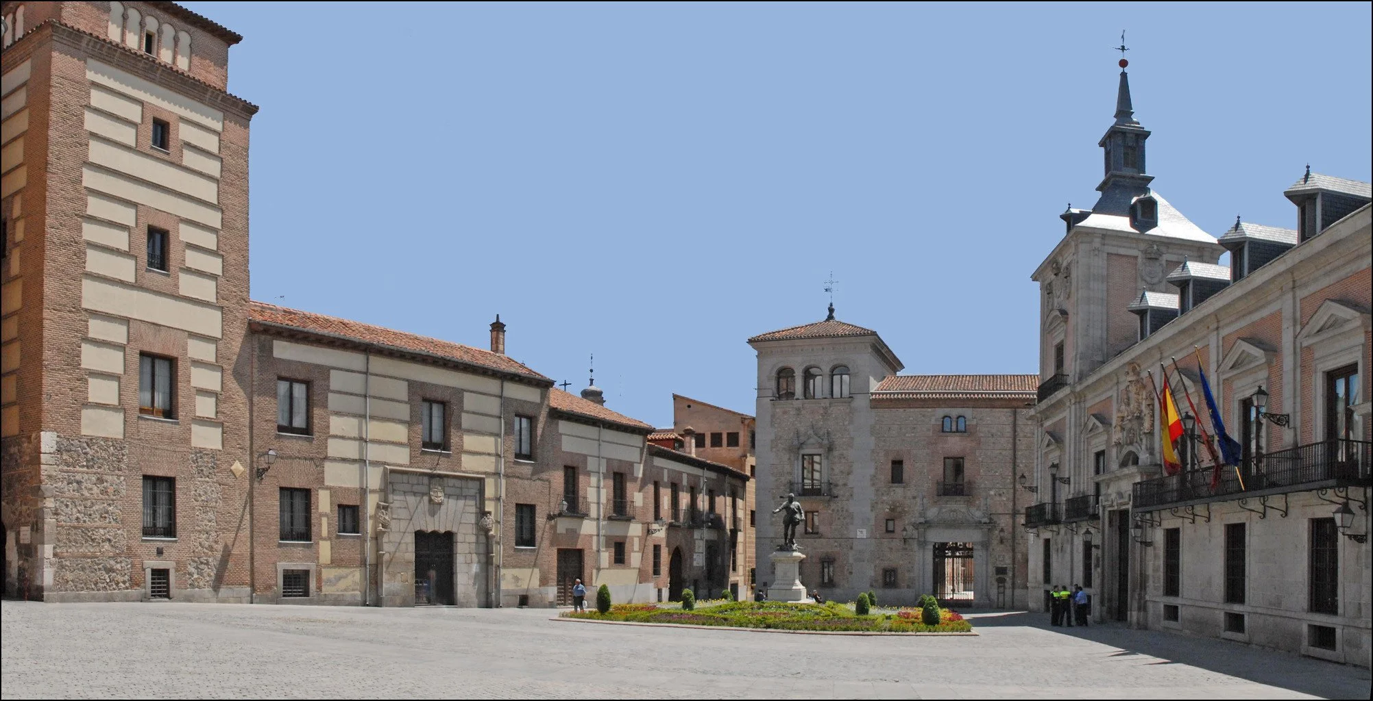 A historic city square with old stone and brick buildings, a statue in a landscaped garden, and flags of Spain and the European Union hanging from a building.
