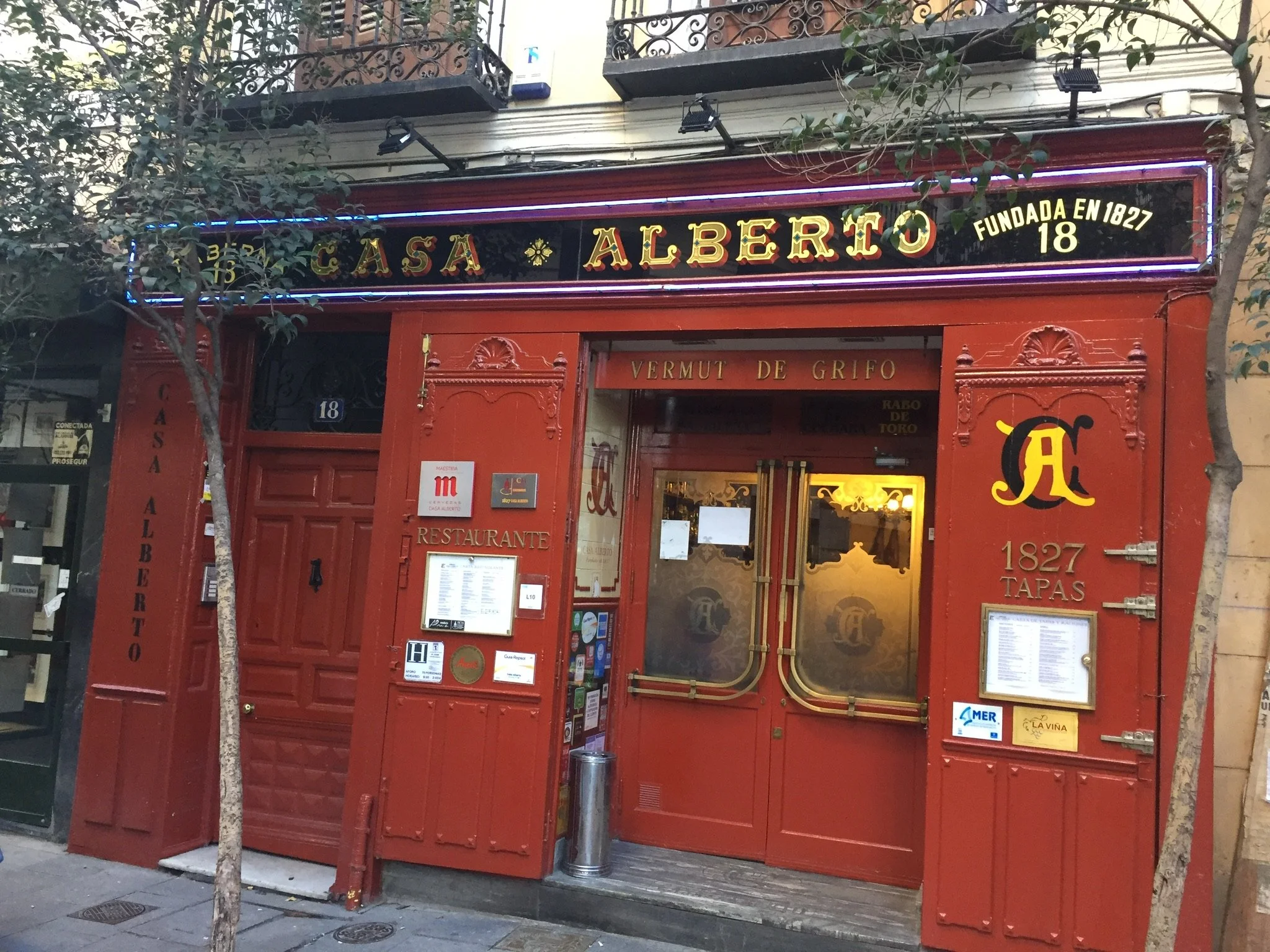Exterior of Casa Alberto restaurant with red facade, glass door, illuminated signboards, and two small trees on the sidewalk.