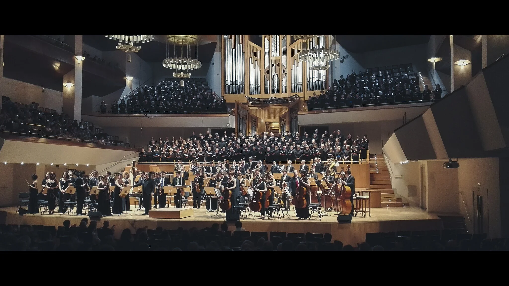 Orchestra performing in a concert hall with a large choir behind them and an organ at the center.