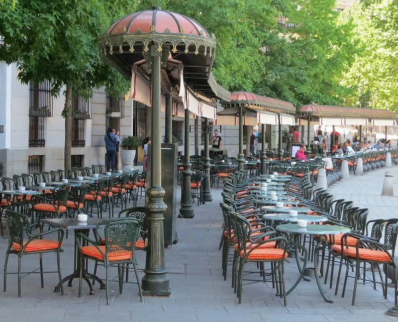 Outdoor café with rows of black chairs and green tables, under large striped umbrellas, near a building with barred windows, with people dining and walking around, surrounded by green trees.