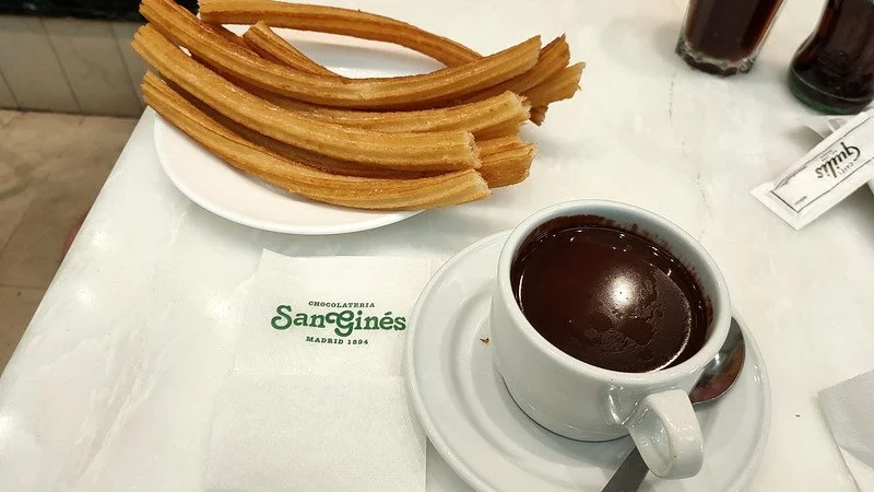 Churros with chocolate dipping sauce on a white plate, hot chocolate in a cup with a spoon, on a table with a napkin that reads 'Sangines'.