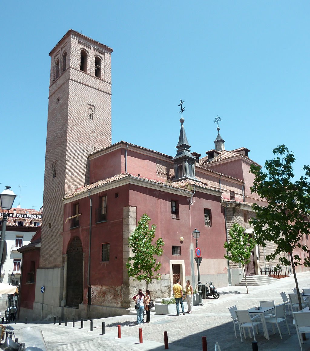 A historic church with a tall bell tower, red walls, and ornate crosses on its roof, situated in a city square with trees, people walking, and outdoor seating under a clear blue sky.