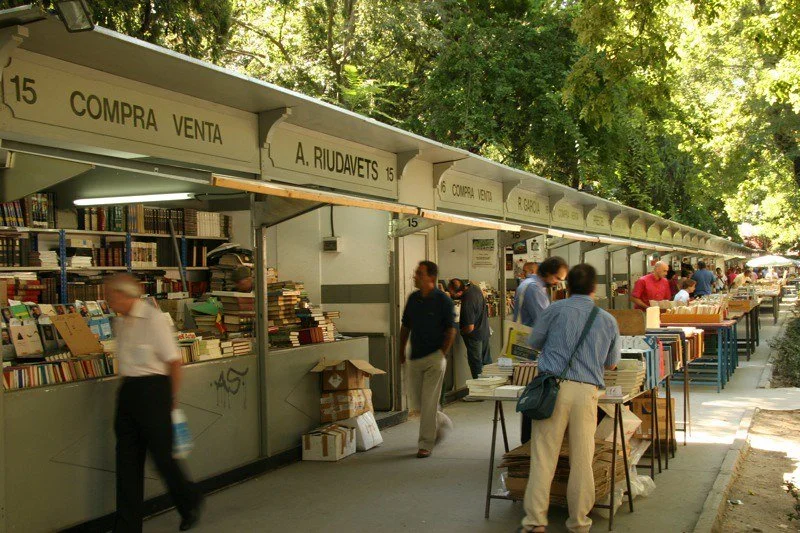 An outdoor book market with booths selling used books, with several people browsing and walking along the row of stalls, surrounded by trees.