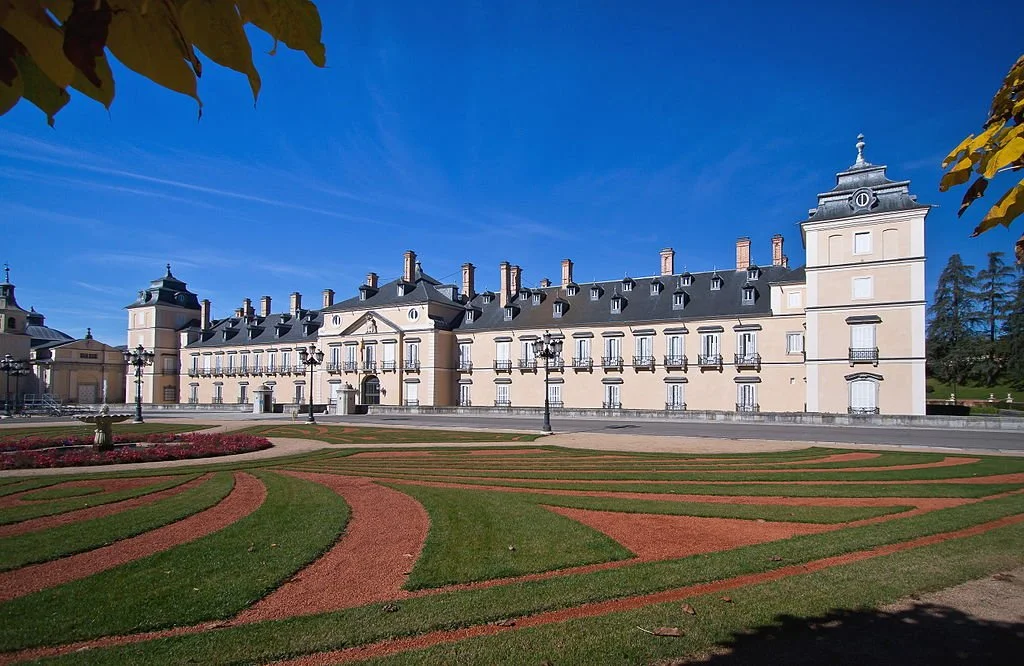 A grand historic castle with white walls and black rooftops, surrounded by a well-maintained garden with colorful flowerbeds and paved walkways, under a bright blue sky.