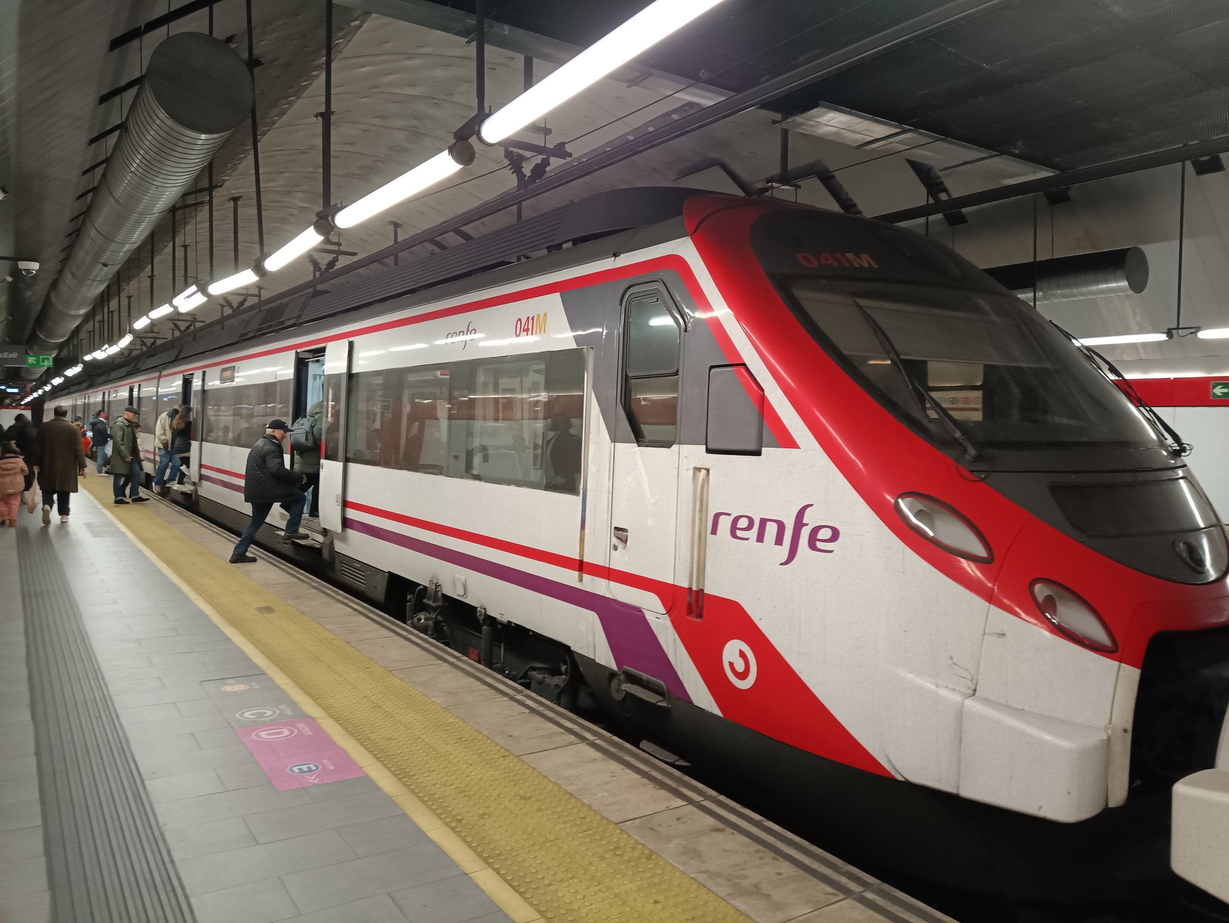 A modern Renfe train at an underground train station with passengers boarding and walking along the platform.