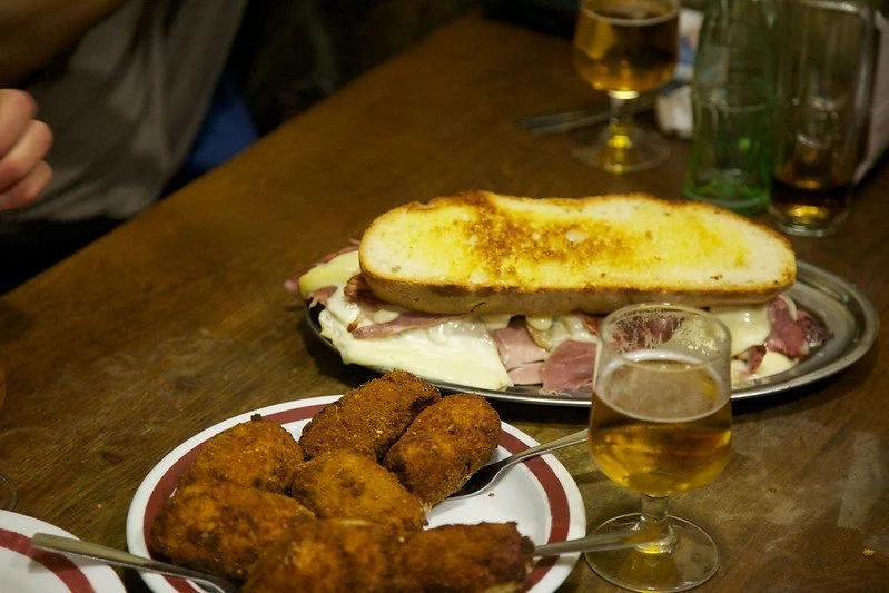 A plate of fried appetizer croquettes, a sandwich with grilled bread, ham, and melted cheese, and glasses of beer on a wooden table.
