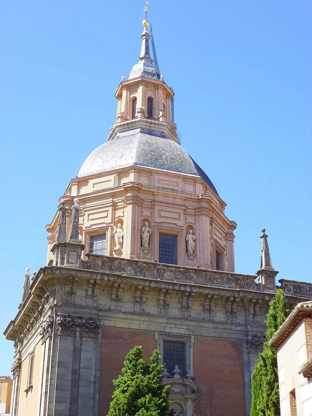 A historic church with a large dome, featuring statues and intricate architectural details, against a clear blue sky. Trees are visible at the bottom.