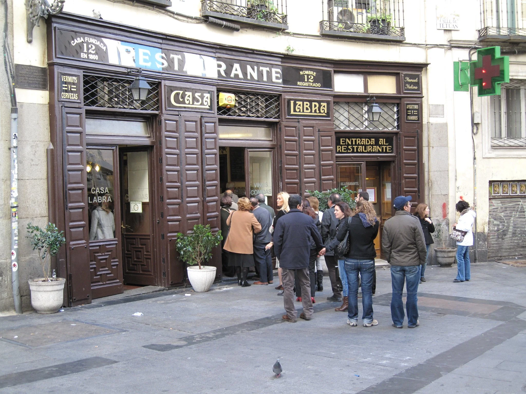 People standing in line outside a restaurant called "Casa Labra" on a city street. The restaurant has a dark brown wooden facade with gold signage, and there is a green pharmacy cross sign on the right side of the building. Potted plants are placed in front of the establishment.