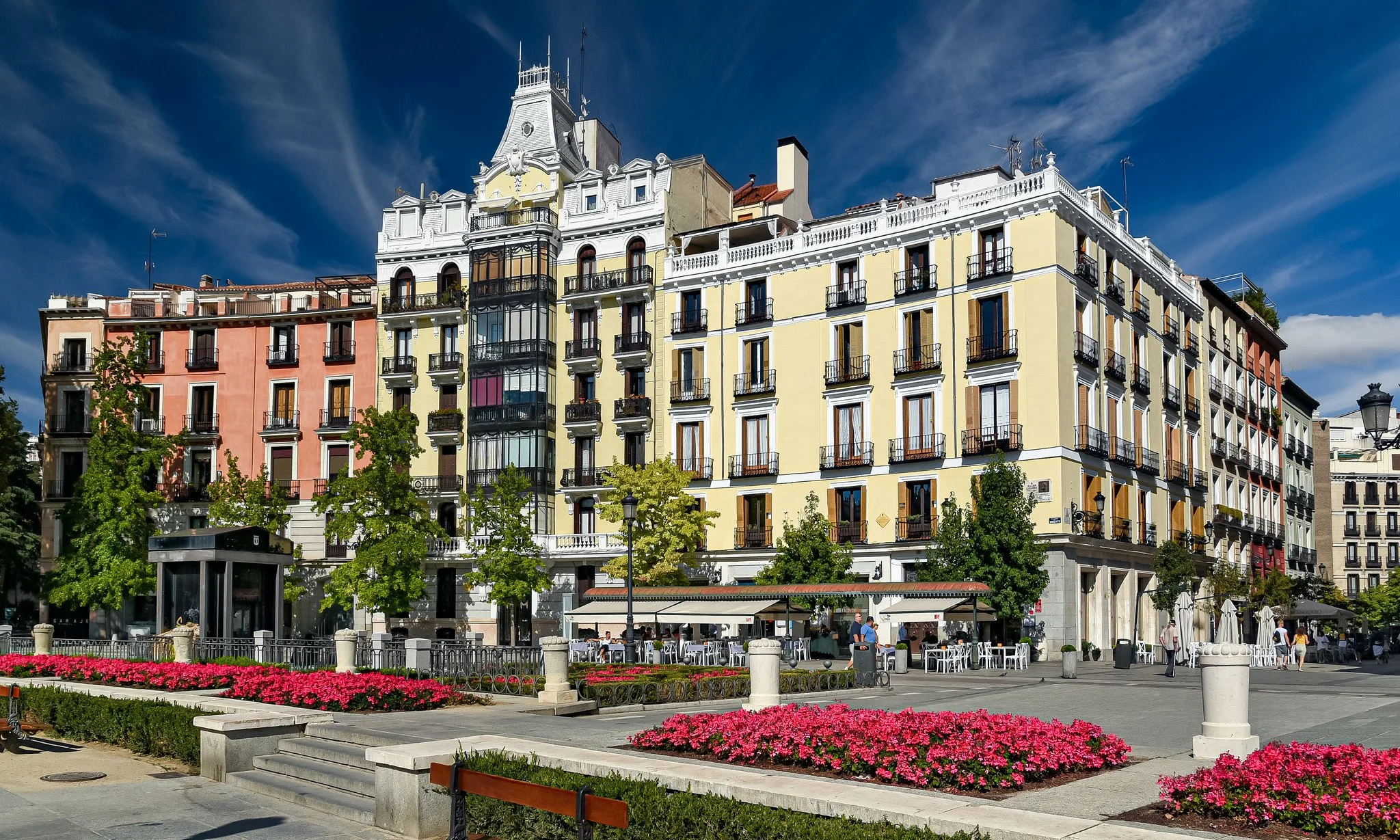 Colorful multi-story buildings with balconies in a city square, green trees, pink flowers, outdoor seating, and a clear blue sky.