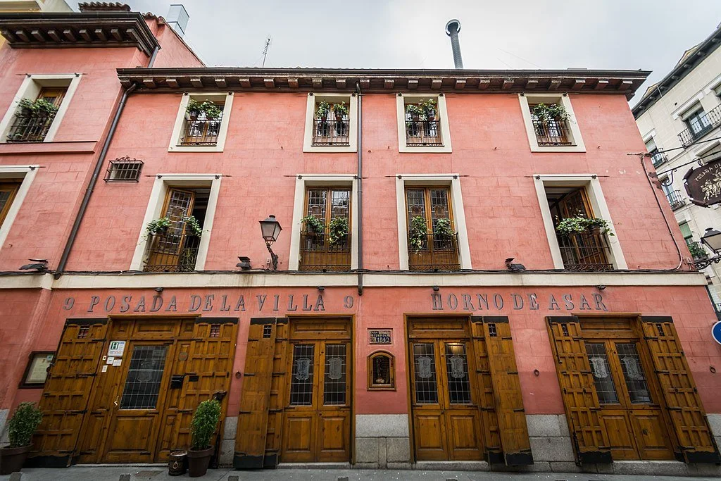 A pink, multi-story building with wooden doors and several windows with flower boxes. Signage indicates it is a restaurant and guesthouse named "Posada De La Villa 9 Horno De Asar."