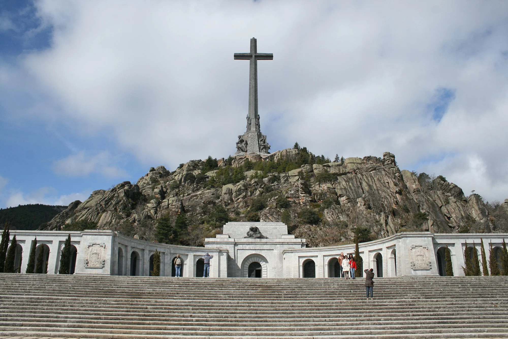 A large stone cross monument atop a rocky hill with a wide staircase and a white colonnade in front, with several people taking photos, under a partly cloudy sky.