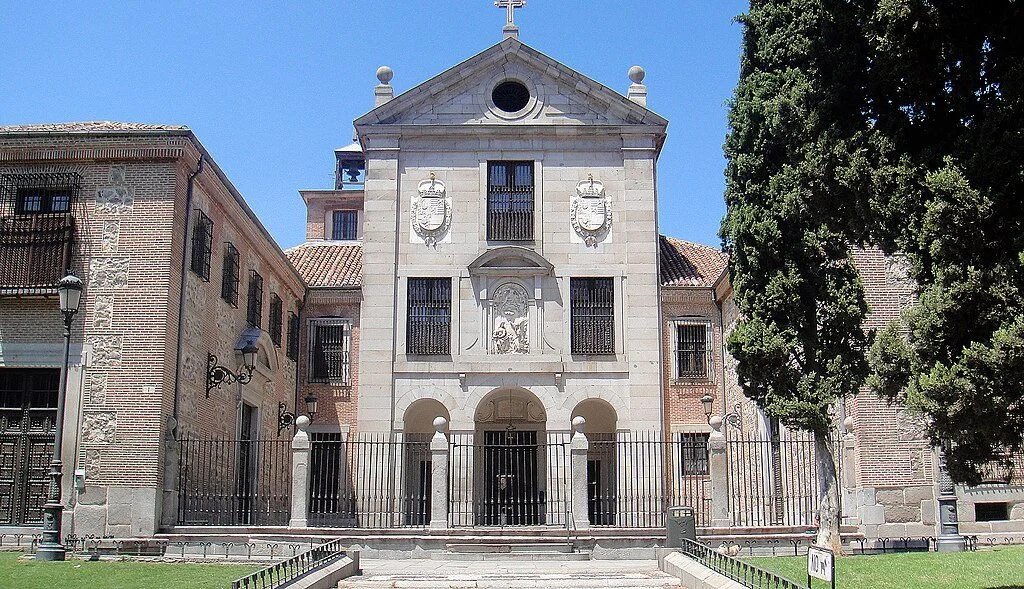 A historic church with a stone facade, decorative shields, and a cross on top, surrounded by a black iron fence and trees.