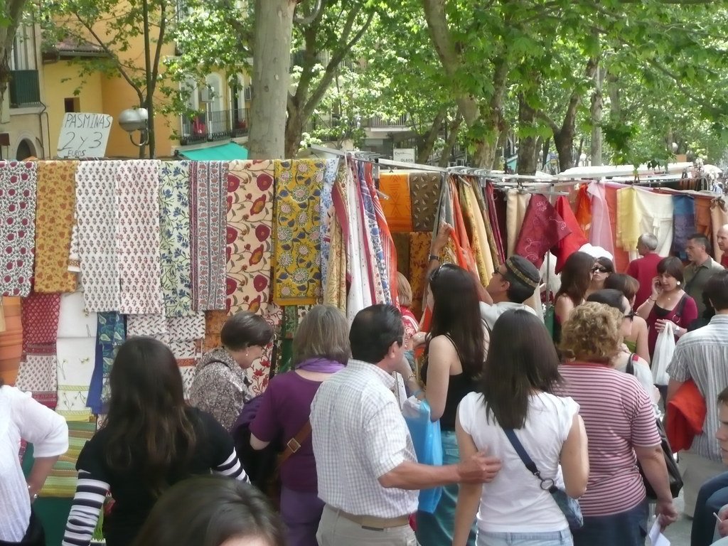 Crowd of people browsing fabrics at an outdoor market stall with colorful textiles hanging from a line, surrounded by green trees.