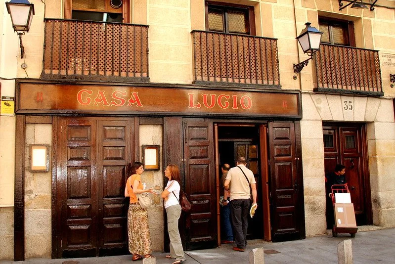 People standing outside a restaurant named Casa Lucio with a dark wooden exterior door and a sign above it. The building has balconies and street lamps.