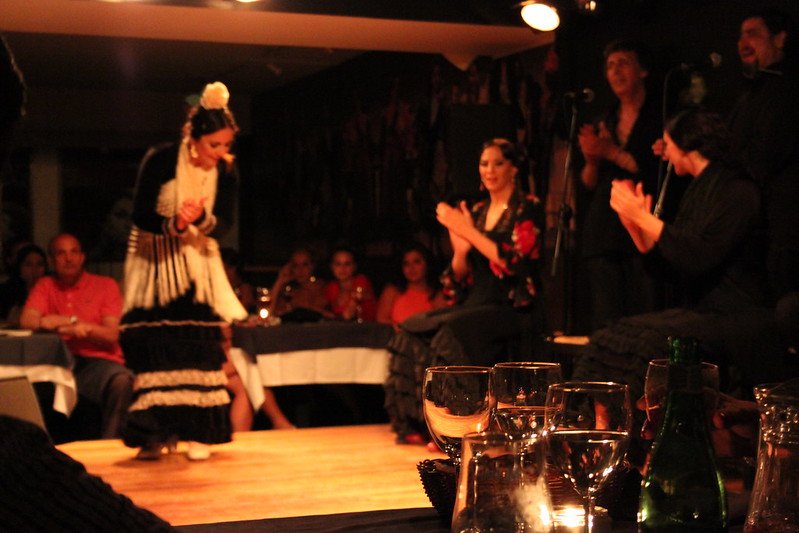A Flamenco performance at a dimly lit venue with a dancer bowing on stage, surrounded by three seated women clapping, and an audience in the background.