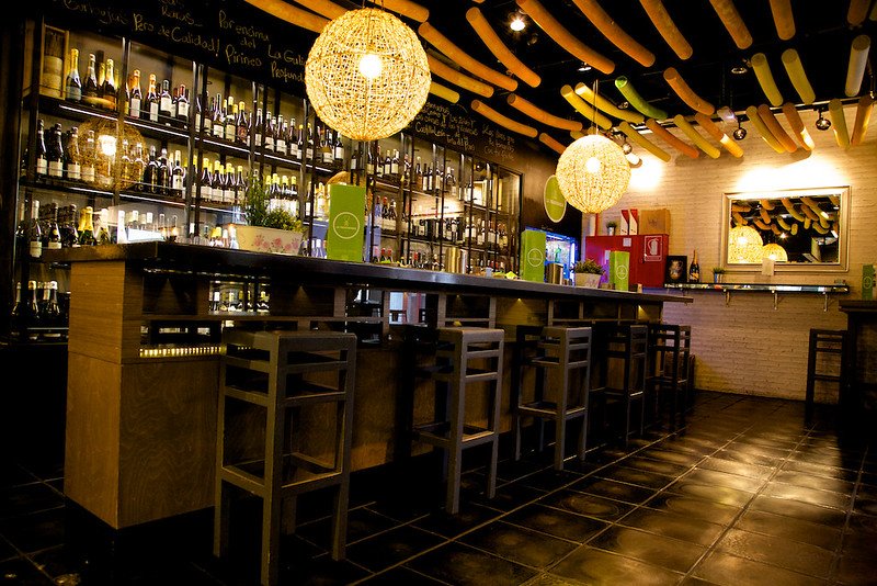 Interior of a bar with a long wooden counter, bar stools, shelves stocked with bottles, and pendant lights with woven shades.