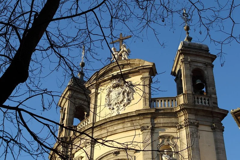Close-up of an ornate church or cathedral with a cross on top, decorated with sculptures and architectural details, against a clear blue sky, with tree branches in the foreground.