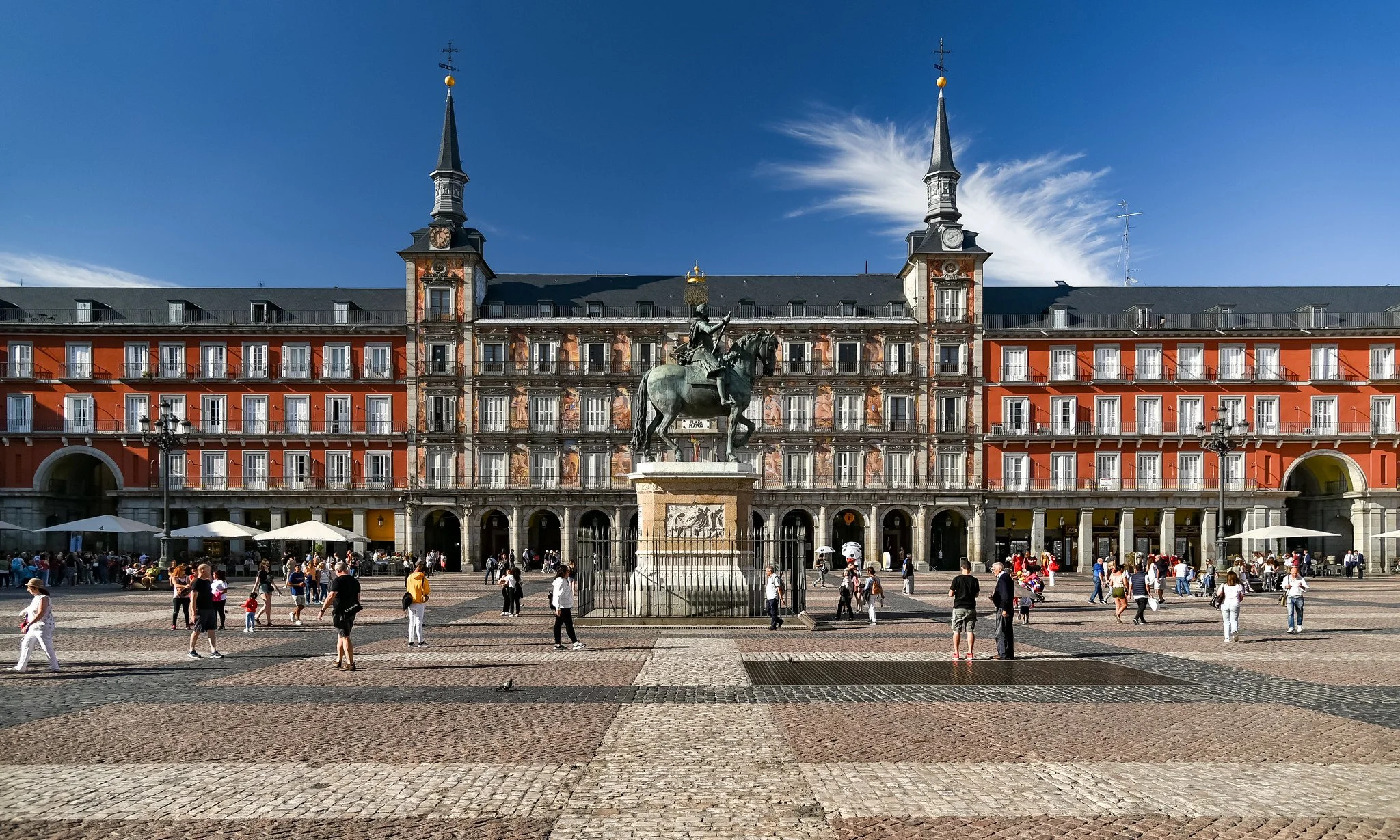 A large public square with a statue of a person on a horse in front of a historic building with turrets, arched windows, and balconies, under a bright blue sky.